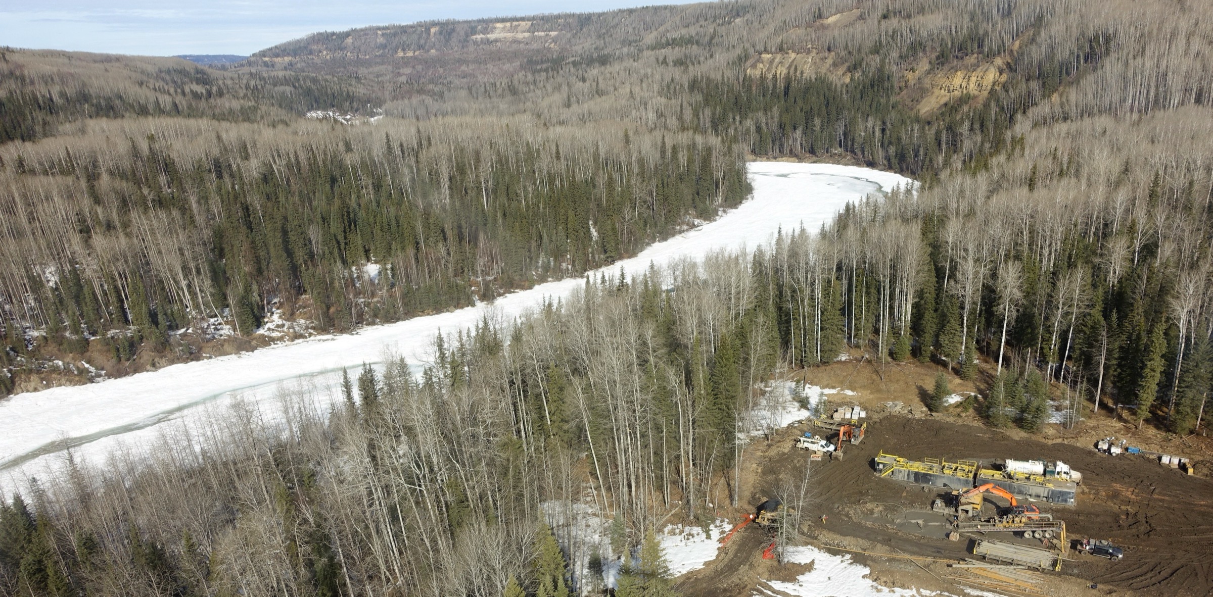 Aerial view of a frozen river valley in boreal forest, with a small drilling spread set on one bank and no cleared right of way visible across the valley